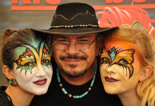 Mark Reid posing between two women he has painted masks on with a butterfly sort of feel. One in green and the other yellow 